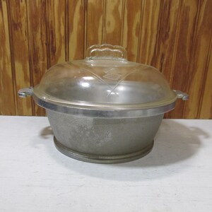 May include: A vintage, round, aluminum cooking pot with a clear glass lid. The pot has two side handles and a clear glass handle on the lid. The pot is sitting on a white surface with a wooden background.