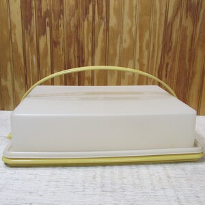 May include: A rectangular, off-white plastic cake carrier with a yellow base and handle. The carrier has a clear lid and a yellow arched handle for easy carrying. The design is simple and functional, suitable for transporting baked goods.