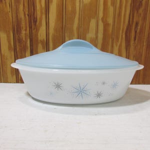 May include: A white oval casserole dish with a blue lid and a starburst pattern. The dish is made of glass and has a vintage look.