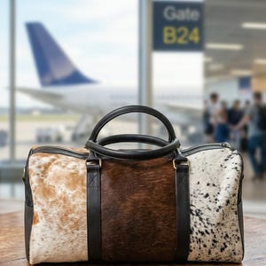 May include: A tri-color travel bag with a cowhide pattern. The bag features a combination of brown, tan, and white spotted sections. It has black leather handles and trim, and a zipper closure. The bag is on a wooden surface.