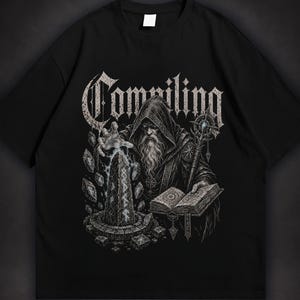Op de afbeelding: Een zwart t-shirt met een grafische print van een tovenaar met een kap en een lange baard, die een staf en een open boek vasthoudt. De tovenaar spreekt een spreuk uit over een stenen obelisk met runen. Het woord Compiling staat erboven.