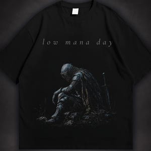 Dark fantasy t-shirt design featuring an exhausted medieval knight sitting with his head lowered beside a sword, with small text reading low mana day above the artwork.