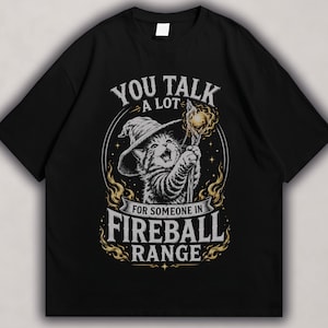 Wizard kitten wearing a floppy hat, holding a magical staff with orange fireball accents and bold “Fireball Range” fantasy text on a black t-shirt design.