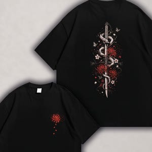 Può includere: T-shirt neri con design grafico. Una maglietta presenta un fiore rosso sul davanti. L'altra ha un design dettagliato sul retro, con una spada, un serpente, fiori rossi e farfalle.