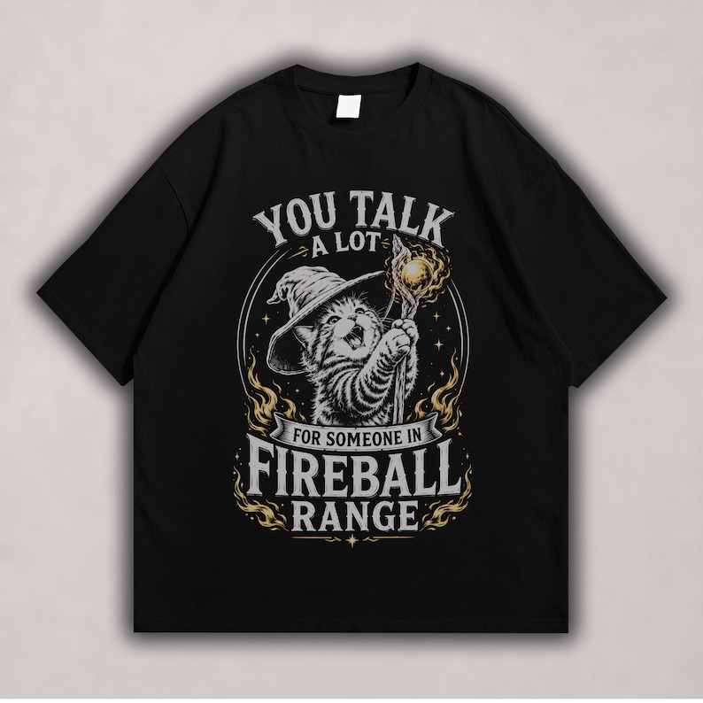 Wizard kitten wearing a floppy hat, holding a magical staff with orange fireball accents and bold “Fireball Range” fantasy text on a black t-shirt design.