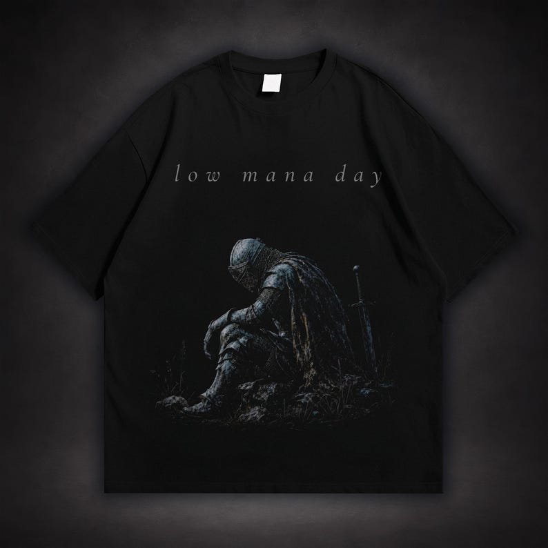 Dark fantasy t-shirt design featuring an exhausted medieval knight sitting with his head lowered beside a sword, with small text reading low mana day above the artwork.