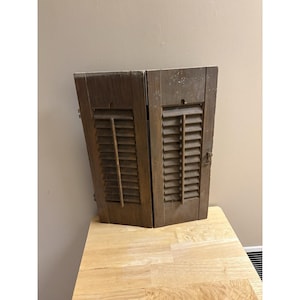Peut inclure: Deux volets en bois patinés avec des lattes horizontales. Les volets sont de couleur marron foncé et semblent vieillis. Ils sont appuyés contre un mur de couleur claire, posés sur une surface en bois.