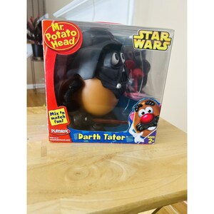 Pode incluir: Um brinquedo Mr. Potato Head em uma caixa de plástico transparente. O brinquedo é Darth Vader de Star Wars. A caixa tem o logotipo de Star Wars e as palavras "Darth Tater". A caixa é vermelha e azul.