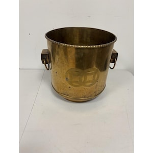 May include: A vintage brass bucket with a round shape and two handles. The bucket has a decorative etched design on the front and a beaded rim. The brass has a warm, aged patina.
