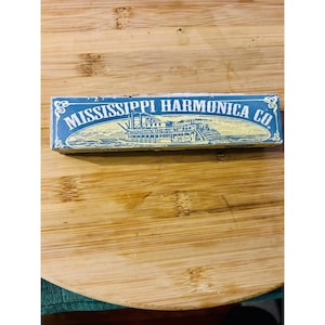 Puede incluir: Una caja azul con las palabras "MISSISSIPPI HARMONICA CO." en letras blancas. La caja presenta una ilustración de un barco de vapor en amarillo y azul. La caja está sobre una superficie de madera.