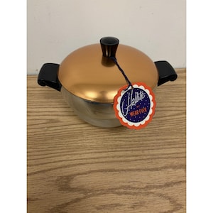 Puede incluir: Una olla de metal color cobre con tapa a juego, que presenta dos asas negras y un pomo negro en la parte superior. Una etiqueta de borde festoneado está unida al pomo con un cordel oscuro, mostrando el texto Hallite by WEAR-EVER en blanco y naranja.