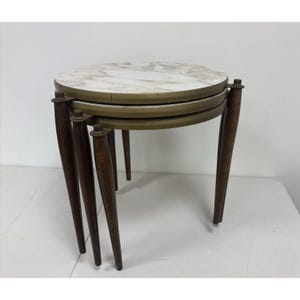 May include: Set of three nesting tables with round marble tops and brass-colored rims. The tables have dark brown, tapered wooden legs. The tables are stacked together, with the smallest table at the bottom and the largest at the top.