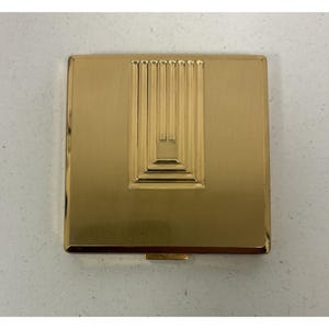 Könnte beinhalten: Ein goldfarbener, quadratischer Kompaktpuder mit einem geometrischen Design auf der Oberseite. Das Design zeigt vertikale Linien und eine abgestufte rechteckige Form. Der Kompaktpuder hat eine glänzende, reflektierende Oberfläche und einen kleinen Verschluss.