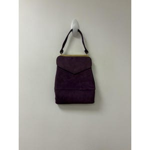 May include: A deep purple suede handbag featuring a structured rectangular body with a pointed flap closure. The bag includes a single matching purple handle attached to a gold-toned metal frame that runs along the top edge of the bag's main compartment.