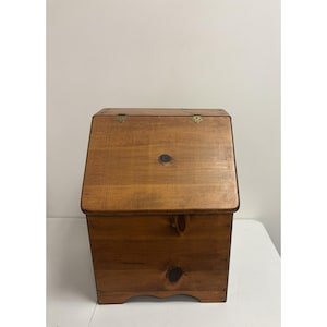 May include: A wooden storage box with a hinged, slanted lid. The box is made of light brown wood with visible knots and brass hinges. The box has a rectangular shape with a slightly curved base.