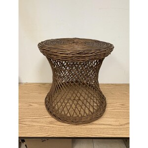 May include: A brown wicker stool featuring a circular top and a flared, hourglass-shaped base constructed from woven rattan strands. The structure displays a crisscross lattice pattern throughout the middle section, providing a stable, open design.