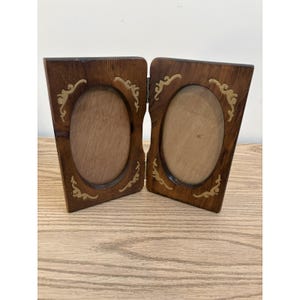 Puede incluir: Un marco de fotos doble plegable hecho de madera marrón oscura con dos aberturas ovaladas. Cada marco presenta detalles decorativos dorados en forma de volutas en las esquinas. Las dos secciones están unidas por una bisagra metálica central.
