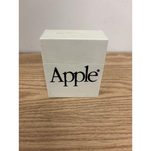 Peut inclure: Une boîte rectangulaire en plastique blanc avec un couvercle, présentant le mot Apple imprimé en police serif noire sur la face avant. Le récipient a une finition mate lisse et une structure carrée propre avec une couture horizontale séparant le couvercle.