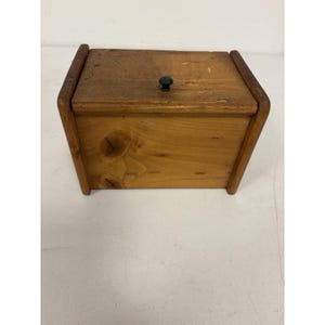May include: A small, rectangular wooden box with a hinged lid. The box is made of light-colored wood with a dark knob on the lid. The corners are rounded, and the box appears to be handmade. The box is likely used for storage.