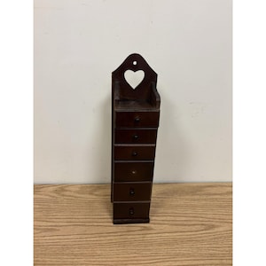 May include: A tall, dark brown wooden wall-mounted organizer featuring six stacked drawers, each equipped with a small black knob. The top section of the unit includes a cutout heart shape and a pre-drilled hole for hanging on a wall surface.