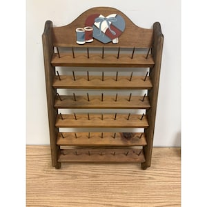 May include: A brown wooden thread rack featuring six horizontal shelves with multiple metal pegs for spool storage. The top arched crest is decorated with a painted heart, two thread spools, and a blue ribbon motif on a light background.
