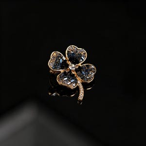 May include: A gold-toned brooch in the shape of a four-leaf clover. The clover's petals are made of faceted, dark blue crystals, with a clear crystal at the center. The stem is adorned with smaller, clear crystals.