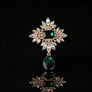 May include: A gold-toned brooch with a large emerald-green rectangular center stone, surrounded by clear, marquise-cut crystals. A teardrop-shaped green stone dangles below. The brooch is set against a black background, reflecting its design.
