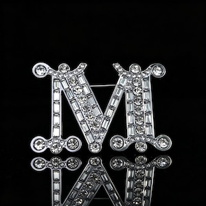 May include: A silver-toned brooch in the shape of the letter "M", encrusted with clear, sparkling crystals. The brooch has a pin on the back for easy attachment. The design is symmetrical and reflects light, creating a dazzling effect.
