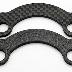 May include: Two black, curved carbon fiber pieces with circular holes. One has a woven pattern, the other a textured appearance. The pieces are likely automotive or industrial components.