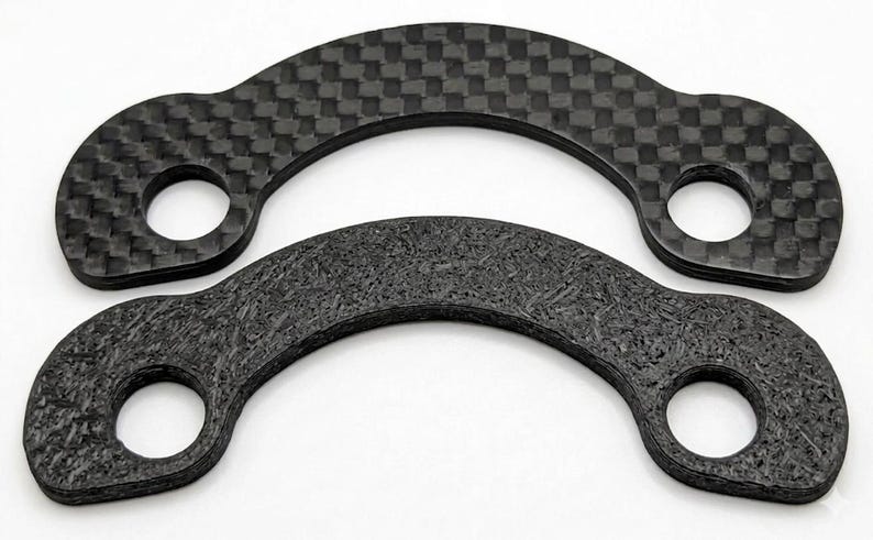 May include: Two black, curved carbon fiber pieces with circular holes. One has a woven pattern, the other a textured appearance. The pieces are likely automotive or industrial components.