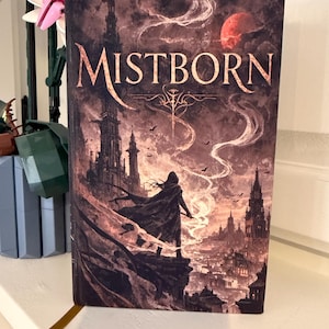 Pode incluir: Um livro de capa dura intitulado "Mistborn" com uma ilustração de fantasia. A capa apresenta uma figura silhueta em um penhasco, uma paisagem urbana e uma lua vermelha. A paleta de cores inclui tons de marrom, vermelho e cinza.
