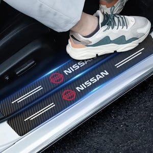 以下が含まれることがあります： 車両用の黒いカーボンファイバー調ドアシルプロテクターのペア。白い日産のロゴとテキスト、赤い円形のエンブレム、各パーツに2本の水平な白いストライプが施されており、車のドアフレームに取り付けられています。