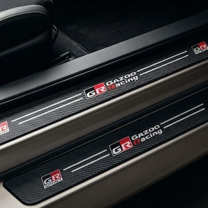 May include: Black carbon fibre textured door sill protectors installed on a vehicle frame. Each piece features the white and red GR Gazoo Racing logo with two horizontal white lines extending across the surface of the rectangular trim plates.