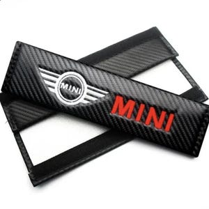 Puede incluir: Dos fundas negras para cinturón de seguridad con un patrón de fibra de carbono. Una funda presenta el logotipo de MINI en blanco y plata, y la palabra "MINI" en rojo. La otra funda tiene la palabra "MINI" en rojo.