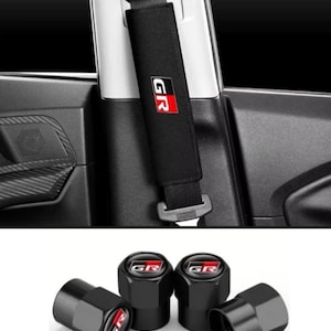 May include: A black seat belt shoulder pad featuring the white and red GR logo, shown installed on a vehicle seat belt. Below are four black hexagonal tyre valve stem caps, each topped with the white and red GR logo on a circular emblem.
