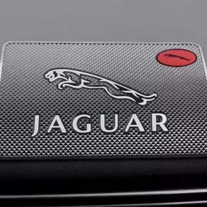 May include: A rectangular, textured silver car emblem featuring the word "JAGUAR" in bold, silver letters. Above the text is a silver jaguar leaping, and a red oval with a jaguar silhouette is in the upper right corner.