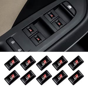 May include: A set of ten rectangular black adhesive decals featuring the red, white, and black GR Gazoo Racing logo. These small stickers are designed to be applied to the window control buttons on a vehicle door panel for a custom appearance.