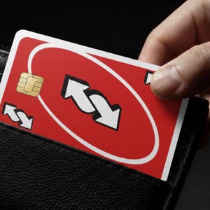 Könnte beinhalten: Eine rot-weiße Uno Reverse-Karte mit einem goldenen Chip wird teilweise in ein schwarzes Lederportemonnaie gesteckt. Die Karte zeigt zwei weiße Pfeile in einem kreisförmigen Design. Das Portemonnaie ist schwarz und hat eine strukturierte Oberfläche.
