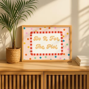 May include: A rectangular framed print featuring a light pink background with multicoloured polka dots. The centre contains a white rectangle with a red and white striped border, displaying the yellow text "Do It For The Plot" in a bold, rounded font.