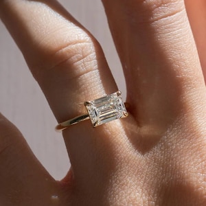 May include: A gold ring featuring a horizontal emerald-cut clear gemstone set in a four-prong setting. The band is thin and polished, showcasing a classic design that highlights the rectangular shape and faceted clarity of the center stone.
