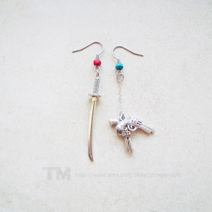 Puede incluir: Un par de pendientes colgantes con dijes de plata. Un pendiente presenta un dije de espada de plata con una cuenta roja en la parte superior. El otro pendiente presenta un dije de pistola de plata con una cuenta turquesa en la parte superior.