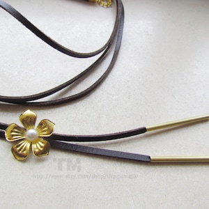 Könnte beinhalten: Eine braune Lederkette mit einem goldenen Blumenanhänger und zwei goldenen Metallketten. Der Blumenanhänger hat eine weiße Perle in der Mitte.