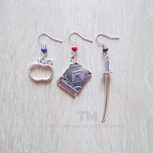 May include: Three silver-toned dangle earrings featuring distinct charms: an apple outline, a rectangular book with a central eye design, and a slender sword. Each earring includes a small colored bead—purple, red, and black—on the hook wire.