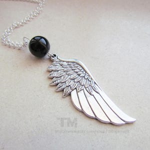 Puede incluir: Un collar con colgante plateado con un diseño de ala detallado. El ala está unida a una cuenta redonda negra y a una cadena plateada. El collar se muestra sobre una superficie gris claro.