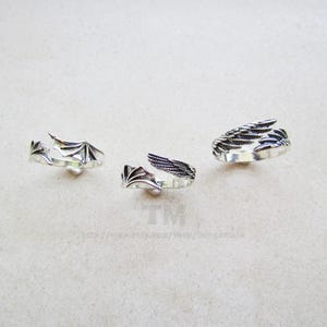 Könnte beinhalten: Drei silberfarbene Ringe mit Flügelmotiven werden vor einem hellen Hintergrund gezeigt. Die Ringe weisen verschiedene Flügelstile auf, darunter fledermausartige Flügel und gefiederte Flügel. Die Ringe sind wahrscheinlich verstellbar, mit offenem Design.