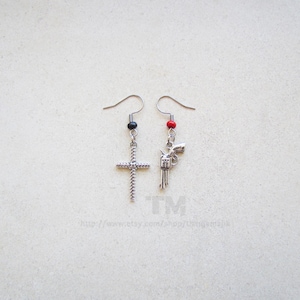May include: A pair of earrings with silver charms. One earring features a silver cross with a black bead, and the other earring features a silver gun with a red bead.