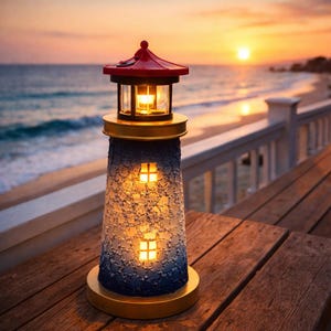 May include: A decorative lighthouse lamp with a red roof, gold accents, and a blue and white textured body. The lamp has two illuminated windows and sits on a wooden surface. The background features a sunset over the ocean.