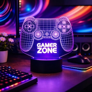 Könnte beinhalten: Eine violette LED-Lampe "Gamer Zone" in Form eines Videospiel-Controllers. Die Lampe hat einen schwarzen Sockel und das Controller-Design ist weiß umrandet. Die Worte "Gamer Zone" sind weiß beleuchtet. Im Hintergrund befinden sich eine Tastatur und ein Monitor.