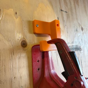 Op de afbeelding: Twee oranje 3D-geprinte gereedschapshouders gemonteerd op een houten oppervlak. Eén houder is boven een rode klem gemonteerd. De houders zijn ontworpen om gereedschap vast te houden. Het hout heeft een natuurlijke nerf.