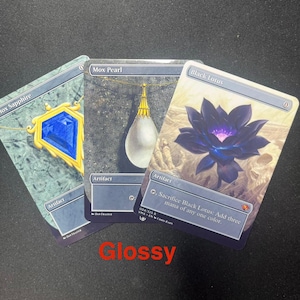 Könnte beinhalten: Drei Sammelkarten: "Mox Sapphire" mit einem blauen Edelstein, "Mox Pearl" mit einem weißen Anhänger und "Black Lotus" mit einer dunkelvioletten Blume. Die Karten sind mit "Artifact" beschriftet und haben Text. Das Wort "Glossy" steht unten.